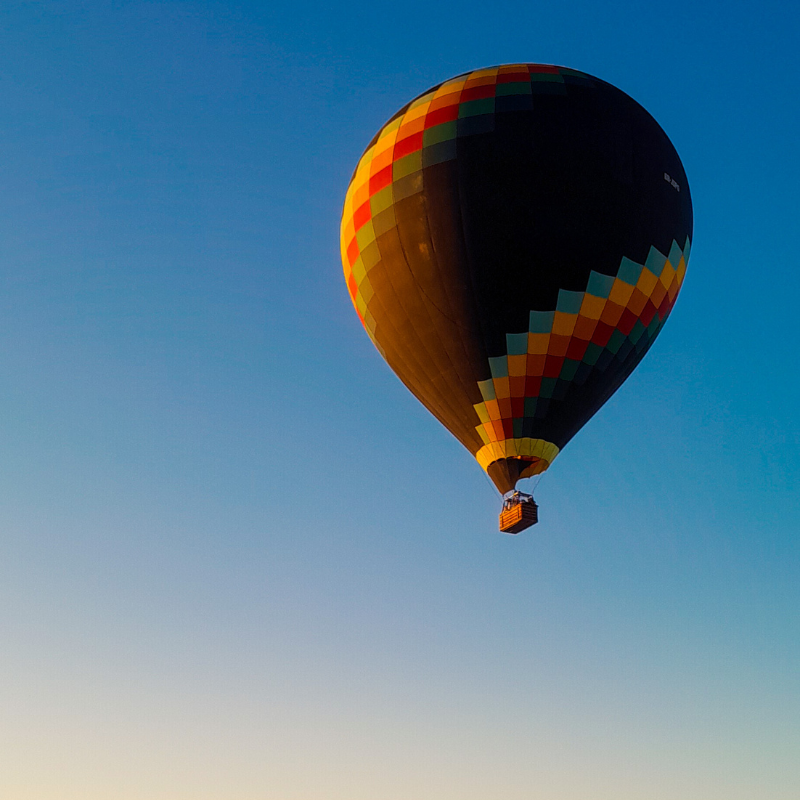 Balão colorido, com desenhos geométricos na parte de cima de varias cores, mesclando com um fundo preto e a cesta pendurada sobrevoa os céus da cidade de Boituva.