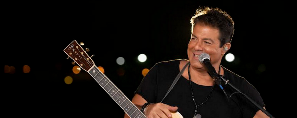Cantor Alex Cohen sorri no palco em frente ao microfone com um violão pendurado no pescoço. Em celebração ao Dia Mundial do Rock, o ator vai se apresentar no Hotel Sheraton São Paulo WTC.