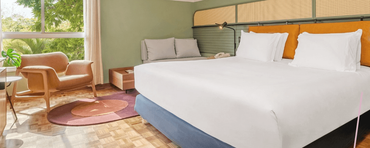 Quarto de hotel com uma cama com lençóis brancos, chão de madeira, uma poltrona em frente à cama e um tapete em formato redondo entre a cama e a poltrona. A cabeceira tem uma luminária e um pequeno sofá ao lado. 