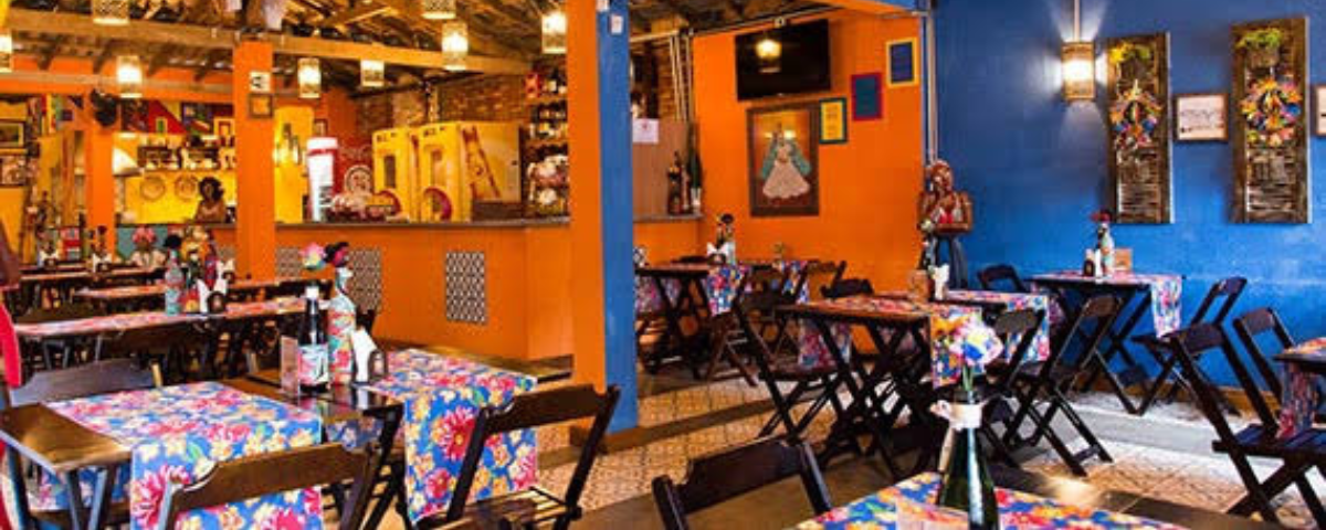 Outro local para comer comida baiana em SP é o Acarajé Botequim. O salão do local tem mesas com toalhas coloridas e as paredes tem as cores azul e laranja. 