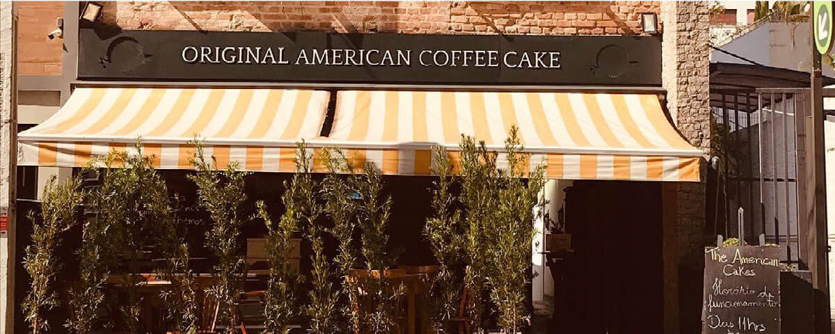 A fachada da The American Cakes tem um toldo listrado nas cores branco e amarelo e o nome da doceria escrito de branco com um fundo marrom. Há também plantas na frente da loja. Por lá você pode encontrar um delicioso cheesecake em SP. 