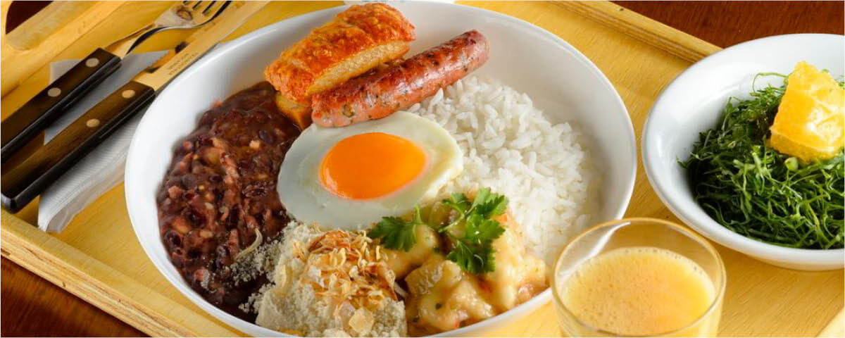 Prato com arroz, feijão, ovo, farofa, linguiça, purê de banana, com um suco e uma porção de couve em tiras servidos ao lado. 