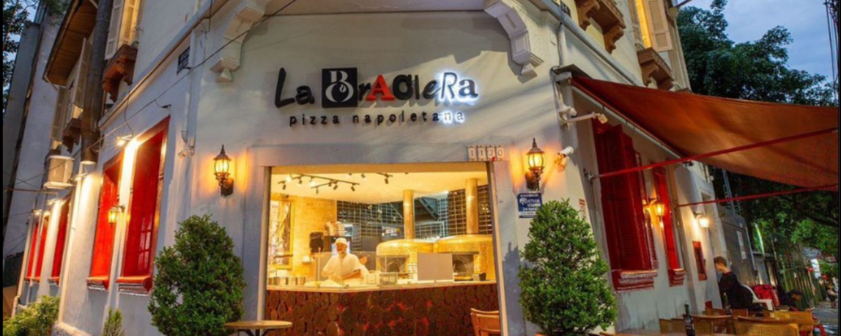 Fachada da pizzaria La Braciera é branca, com janelas vermelhas e o letreiro tem o nome do restaurante nas cores preto e vermelho. 