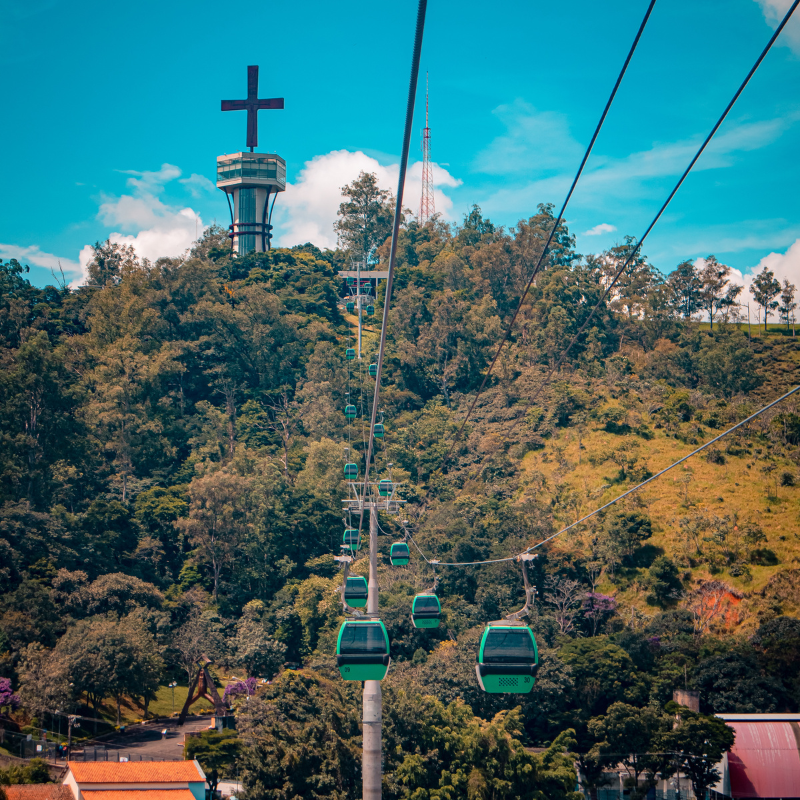 Teleféricos passam por cima da cidade de Aparecida. Ao redor há muitas árvores e os teleféricos são na cor verde. 