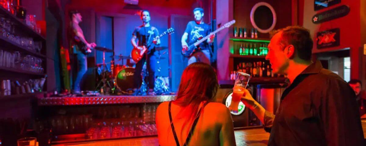 Banda se apresenta no palco de um pub no bairro do Cambuí. Luzes azuis e vermelhas iluminam o local e uma mulher e um homem observam a apresentação. 