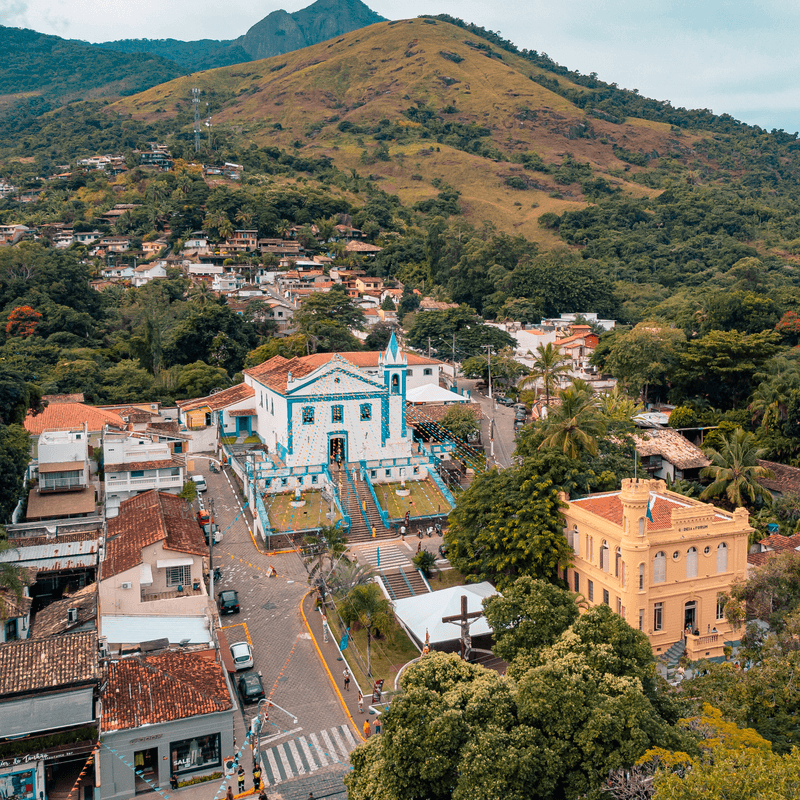 Vista da Igreja de Nossa Senhora D'Ajuda e Bom Sucesso. Ela tem as cores azul e branco, ao redor há algumas casas e ao fundo há montanhas. 