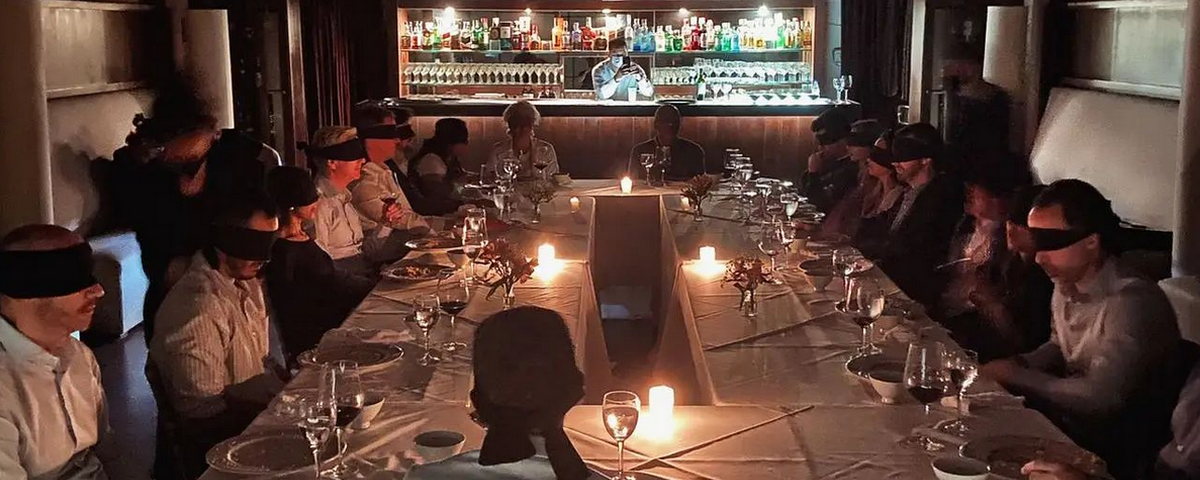 Pessoas jantam usando vendas e o lugar é iluminado apenas com a luz de velas. A experiência é parte do "Jantar no Escuro". 
