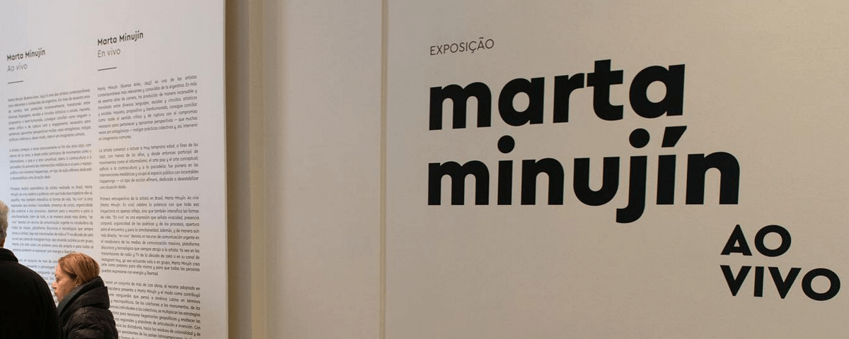 Parede com o nome da exposição em SP "Marta Minujín: Ao Vivo" escrito. Ao lado há um texto falando sobre a mesma. 