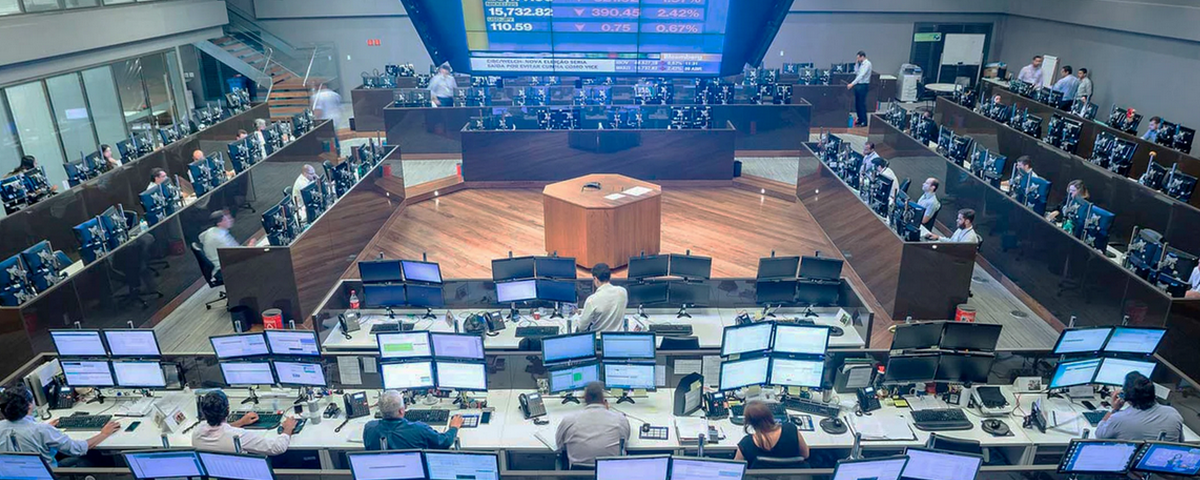 Sala da Bolsa de Valores em São Paulo, com diversos computadores e telas e pessoas sentadas trabalhando. 