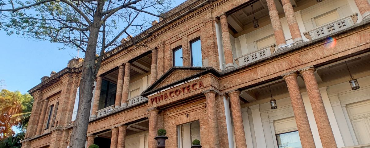 A fachada da Pinacoteca é marrom, com tijolos compondo as paredes e com o nome "Pinacoteca" escrito em vermelho. 