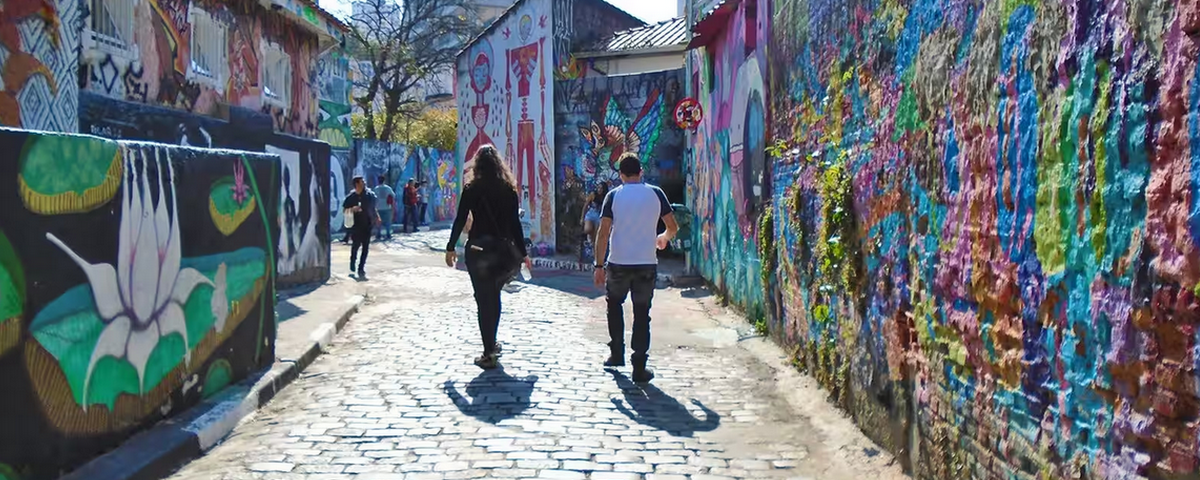 Duas pessoas caminham em rua do Beco do Batman, com diversos grafites desenhados nas paredes ao redor. 