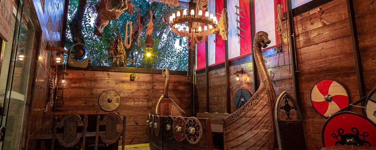 Entrada de taberna, com uma barca de madeira, um lustre e diversos objetos de decoração que remetem a antiguidade. 