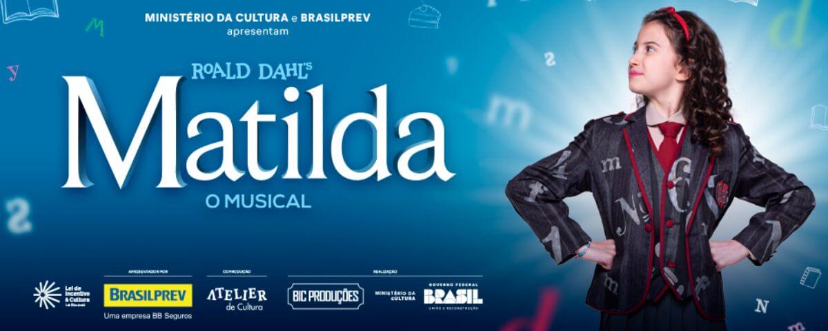 Banner do musical "Matilda" tem as informações do mesmo e uma foto da personagem principal com a mão na cintura do lado direito. 