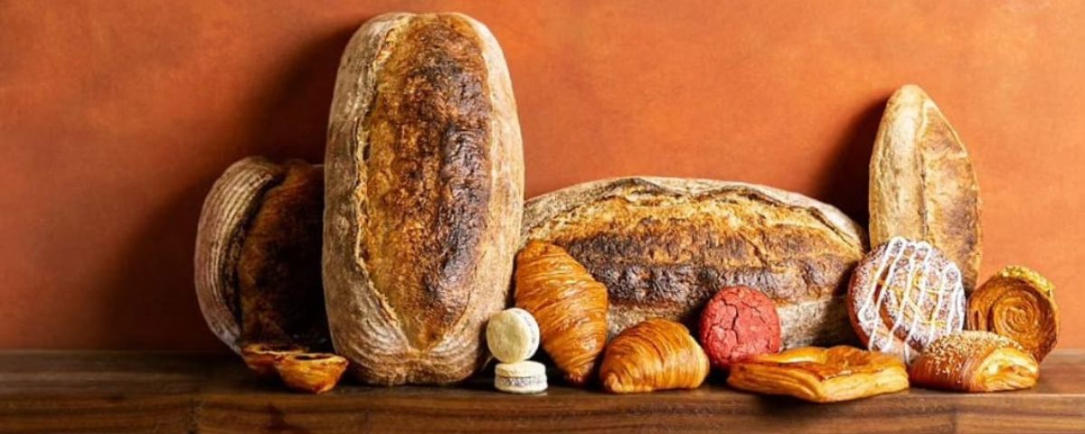 Diversos tipos de quitutes, como pão integral, pão ciabatta, croissaint e macarrons são expostos em prateleira com uma parede laranja ao fundo. 