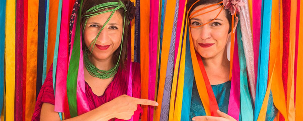 Duas mulheres em meio a diversas fitas coloridas. 