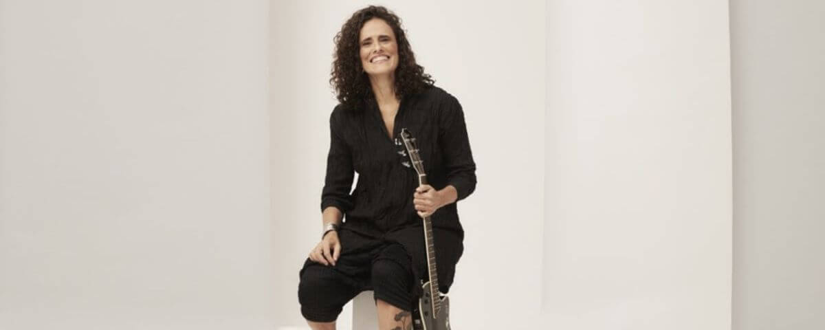 Cantora Zélia Duncan usando uma roupa toda preta e sorrindo enquanto segura um violão. 