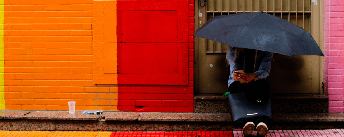 Pessoa sentada em uma calçada com um guarda-chuva cobrindo a cabeça e uma parede pintada de laranja e vermelho ao lado. 