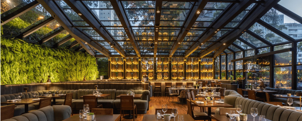 Salão do Must Restaurant é amplo, com móveis em tons de marrom e uma parede viva com plantas do lado esquerdo. 