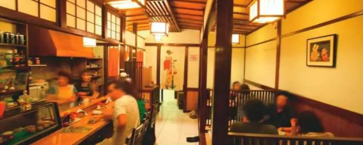 Salão do Restaurante Izakaya Issa, no Bairro da Liberdade, com detalhes de madeira e lugares para sentar no balcão. 