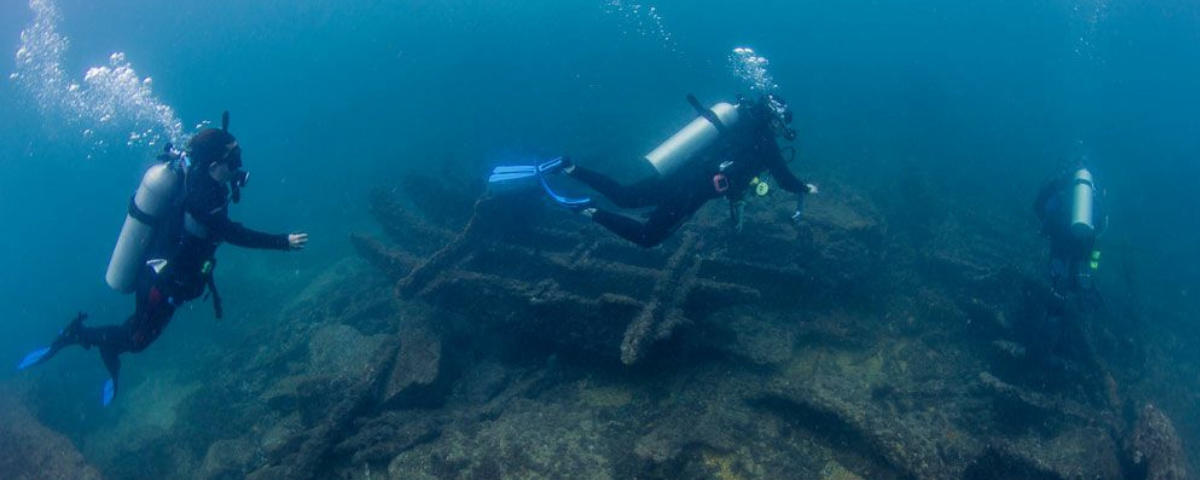 Mergulhadores em baixo d'água passando perto de um navio que naufragou. 